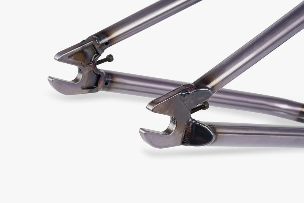 Close-up of raw finish BMX bike frame rear dropouts with integrated chain tensioners, showing welded chainstays and seatstays meeting the axle slot with a bolt for wheel attachment
