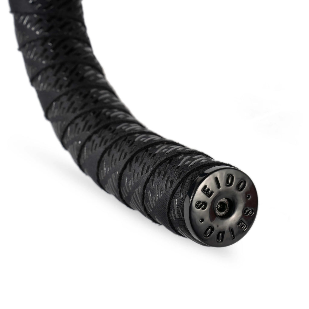 Close-up view of the SEIDO TAB Handlebar Tape, black, highlighting the textured pattern and the glossy black metal SEIDO bar end plug.