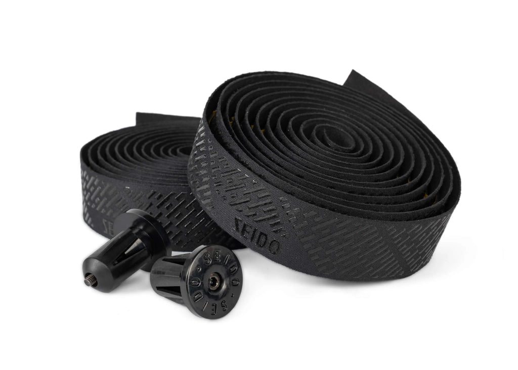 SEIDO TAB Handlebar Tape, black, shown rolled up with the two black bar end plugs disassembled, highlighting the textured grip pattern and SEIDO branding.