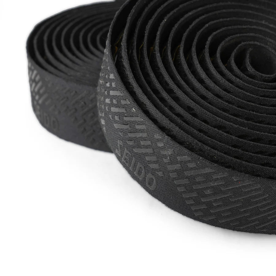 Close-up of the SEIDO TAB Handlebar Tape, black, shown rolled up, highlighting the subtle textured grip pattern and SEIDO branding printed on the tape itself.